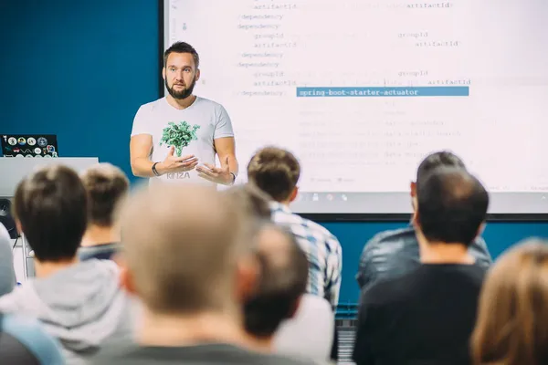 Mikalai talking to a room of engineers, with code on the screen behind him